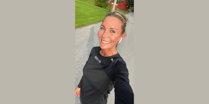 En leende person som tar en selfie utomhus, klädd i en svart träningsjacka och hörlurar, stående på en asfalterad väg med grönt gräs och en röd byggnad i bakgrunden.