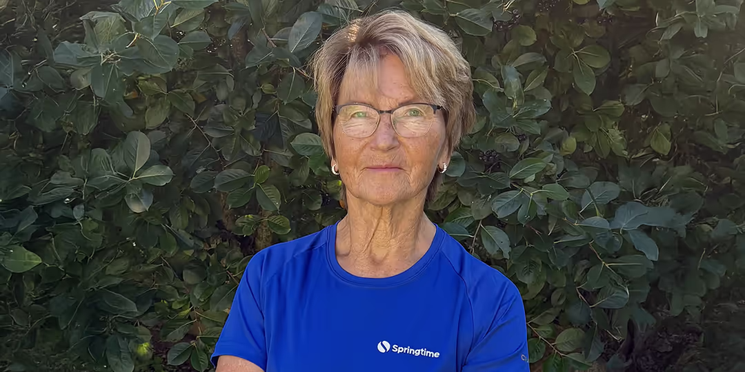 Carina Langemar Äldre kvinna med glasögon och kort ljust hår står framför grönskande buskar. Hon bär en blå t-shirt med logotypen "Springtime" och har armarna i kors.