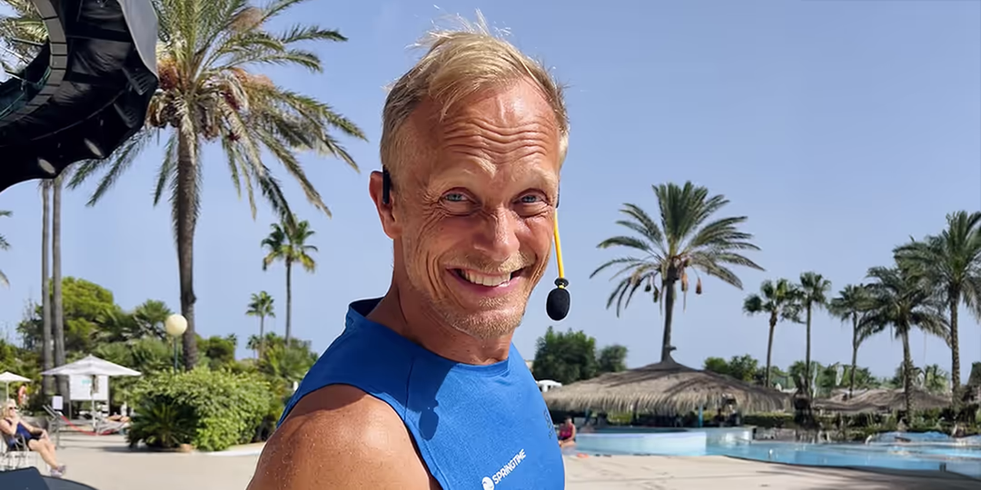 Tobias Karlsson Vältränad man i blått linne med logotypen "Springtime" ler mot kameran. Han bär headset och står utomhus vid en pool med palmer i bakgrunden.