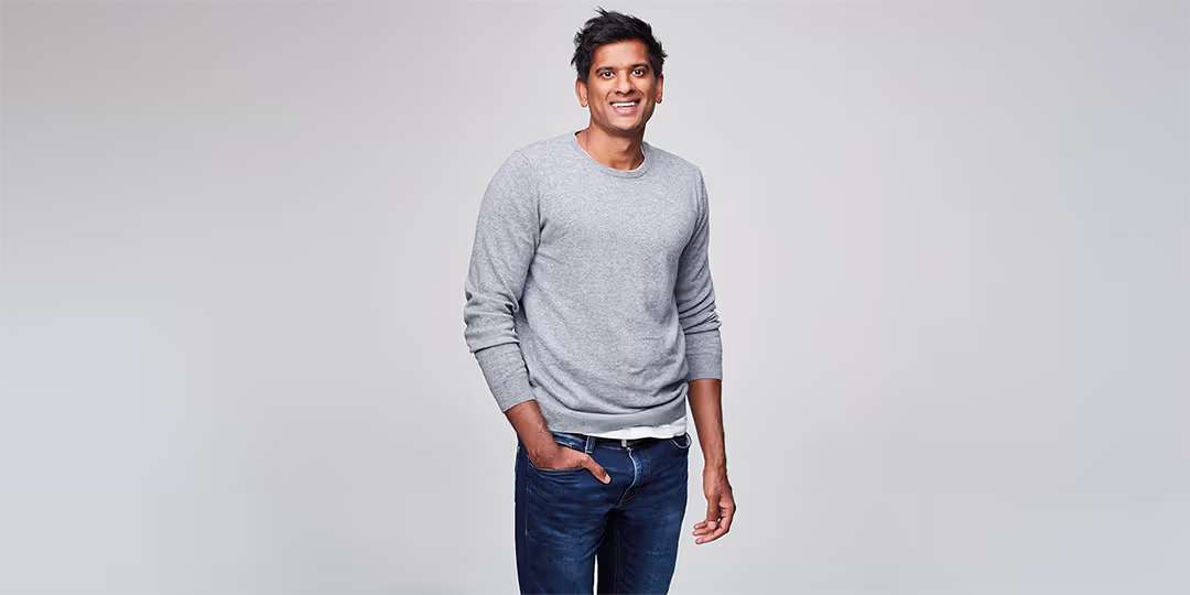 Dr Rangan Chatterjee En leende man i grå tröja och blå jeans som står mot en ljusgrå bakgrund.