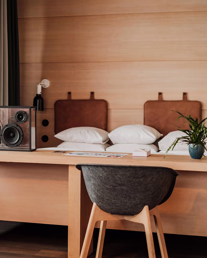 hobo-puff Modernt hotellrum med skrivbord, grå stol, högtalare, växt i kruka och säng med bruna lädergavlar.